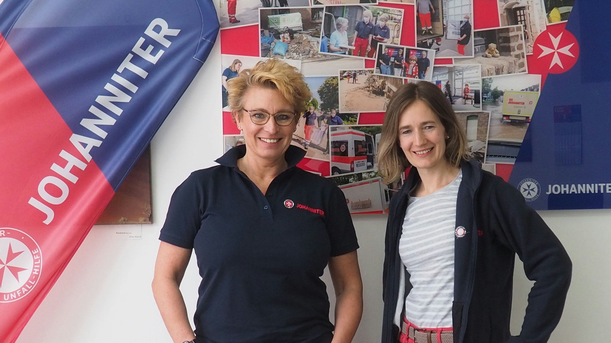 Zwei Frauen stehen vor einer Wand mit Fotos neben einem „Johanniter“-Banner.