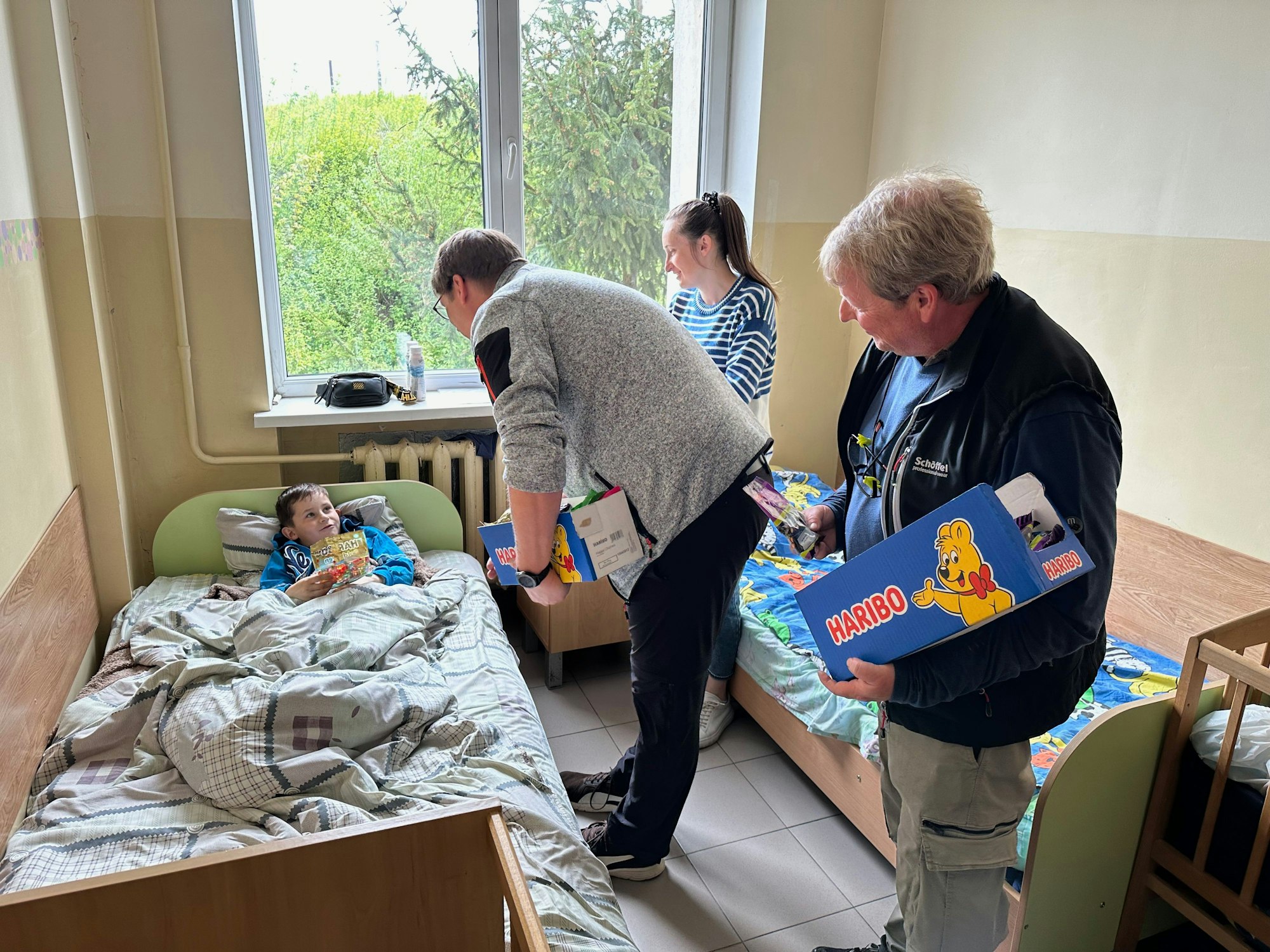 Zwei Männer mit Haribo-Kartons stehen in einem Krankenzimmer, mit einer Frau und einem Jungen, der im Bett mit einer Gummibärchen-Tüte sitzt.