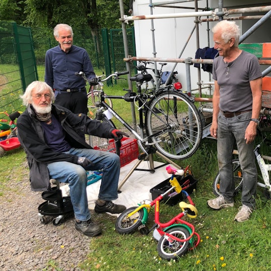 Drei ältere Männer stehen bei einem Fahrrad, das repariert wird, an einem Gerüst.
