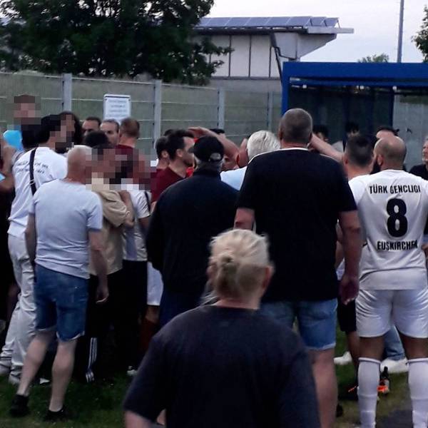 Der Schiedsrichter und ein Assistent stehen am Zaun des Erftstadions, vor ihnen eine aufgebrachte Menge und Menschen, die sie trennen.