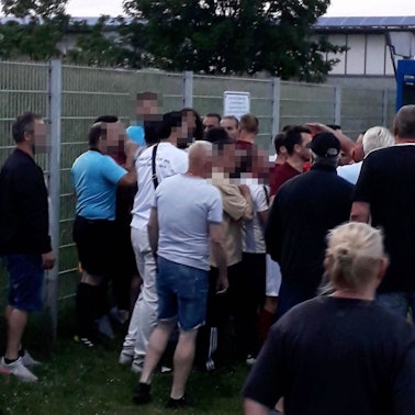 Der Schiedsrichter und ein Assistent stehen am Zaun des Erftstadions, vor ihnen eine aufgebrachte Menge und Menschen, die sie trennen.