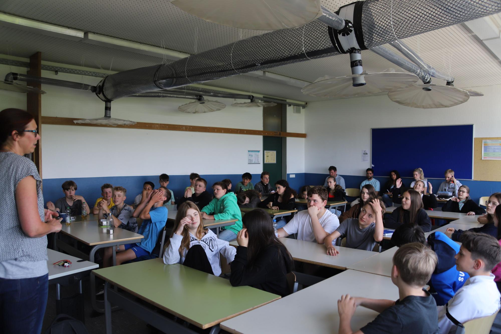 Eine Lehrerin steht vor einer Schulkasse. Die Schüler und Schülerinnen sitzen an Tischen.