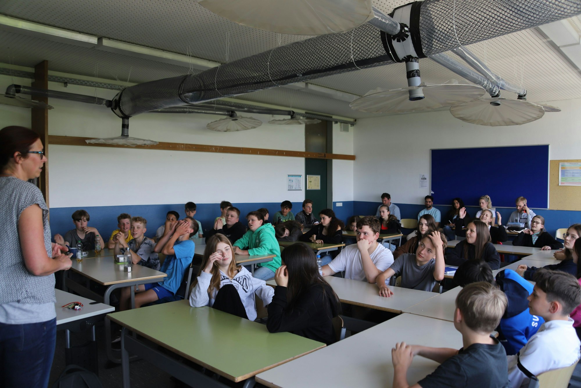 Eine Lehrerin steht vor einer Schulkasse. Die Schüler und Schülerinnen sitzen an Tischen.