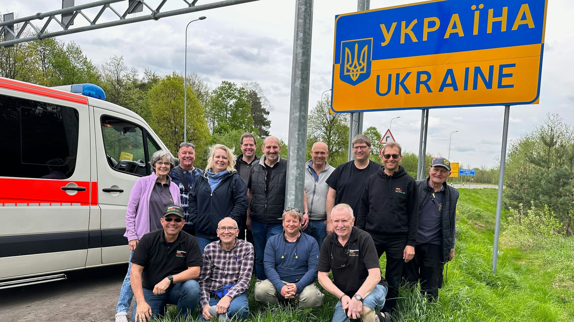 Helfer und Helferinnen stehen zusammen vor einem Grenzschild zur Ukraine neben einem Krankenwagen.