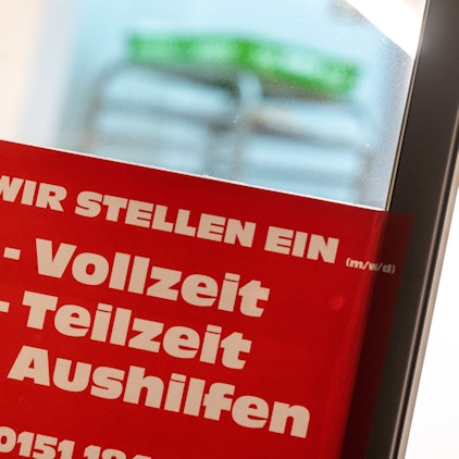 Ein rotes Schild mit der Aufschrift ·„Wir stellen ein - Vollzeit, Teilzeit, Aushilfen“ hängt an einer Eingangstür.
