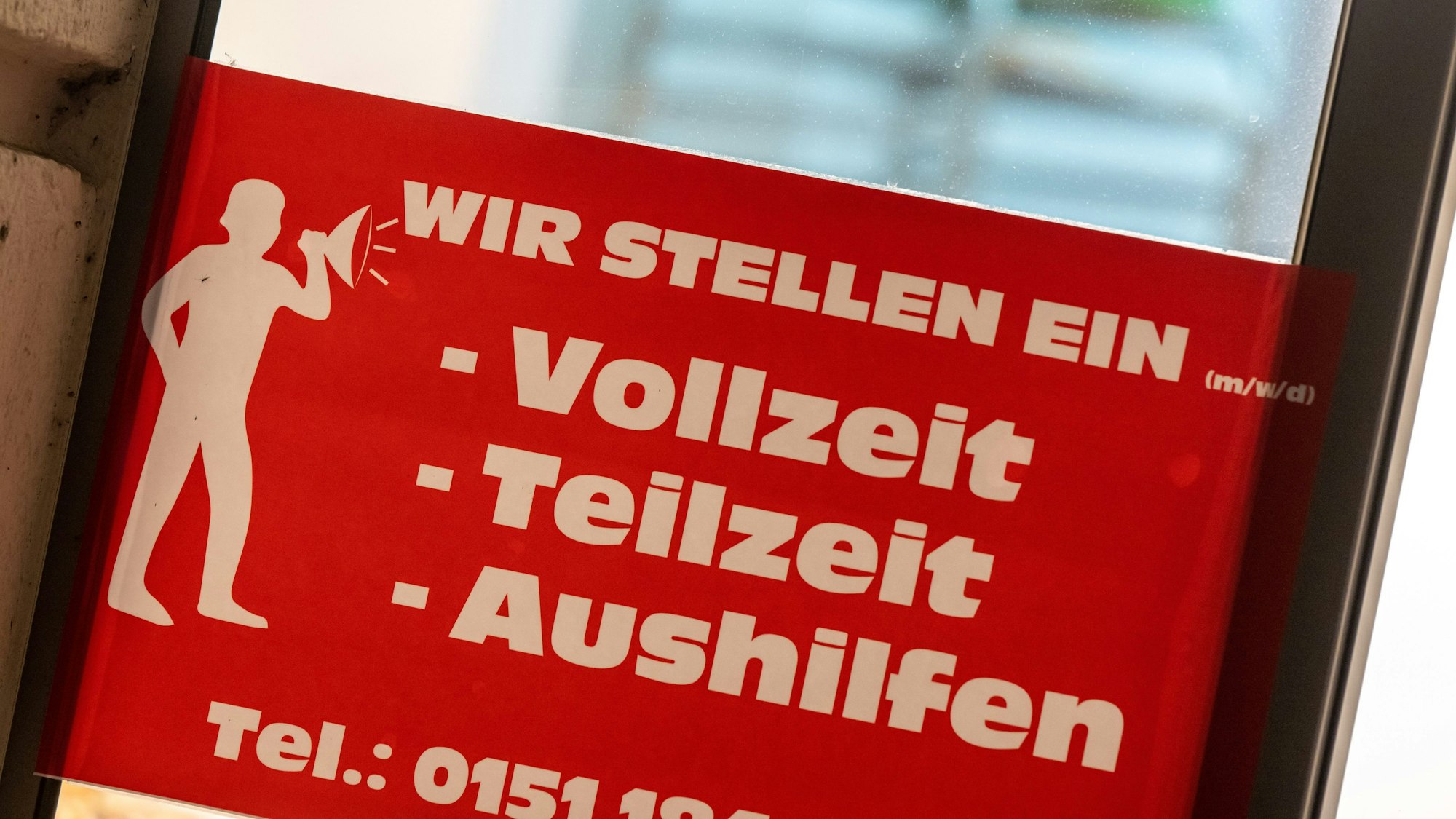 Ein rotes Schild mit der Aufschrift ·„Wir stellen ein - Vollzeit, Teilzeit, Aushilfen“ hängt an einer Eingangstür.