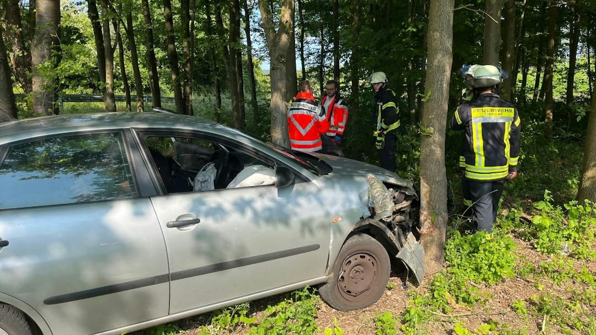 Ein beschädigtes Auto steht vor einem Baum. Feuerwehrleute sind im Einsatz.