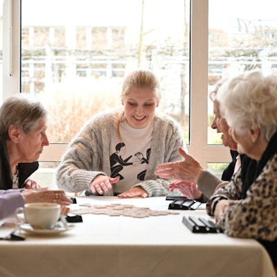 Eine junge Frau sitzt mit vier Bewohnerinnen einer Seniorenresidenz am Tisch und spielt mit ihnen ein Gesellschaftsspiel.