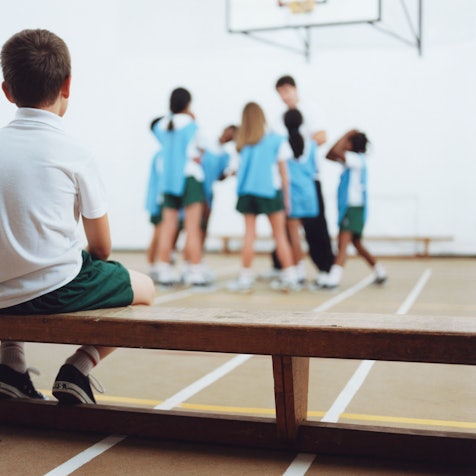 Ein kleiner junge sitzt alleine auf der Bank im Sportunterricht.