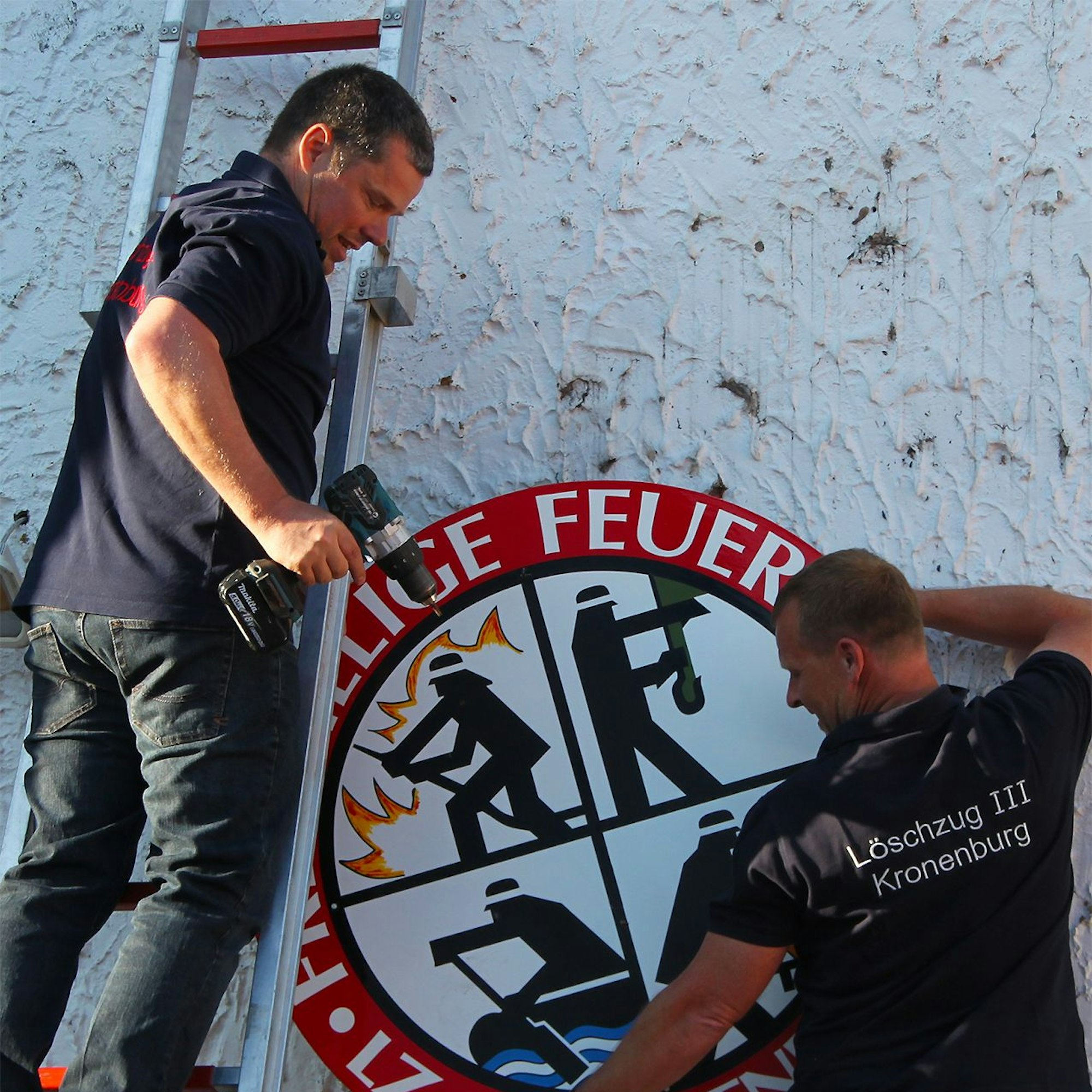 Das Bild zeigt zwei Männer, die auf Leitern stehen und ein Feuerwehr-Schild von einer Fassade abnehmen.