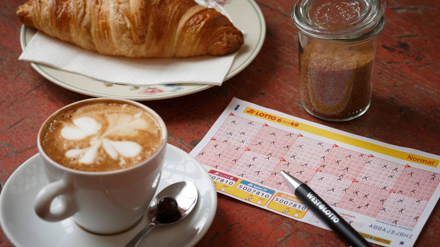 Lotto 6aus49 Spielschein auf Tisch neben Kaffeetasse und Croissant.