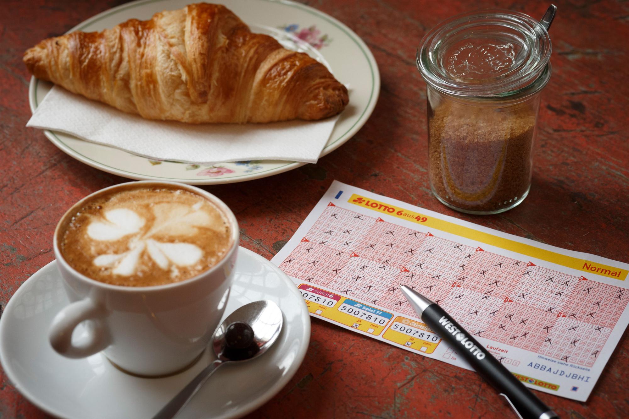 Lotto 6aus49 Spielschein auf Tisch neben Kaffeetasse und Croissant.