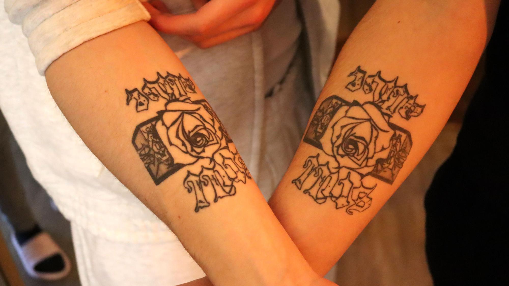 Liaze und MTJ zeigen ihre Arme, auf die sie den Namen des Labels Still Alive und das Logo, eine Rose, die aus Betonbauten wächst, tätowiert haben.