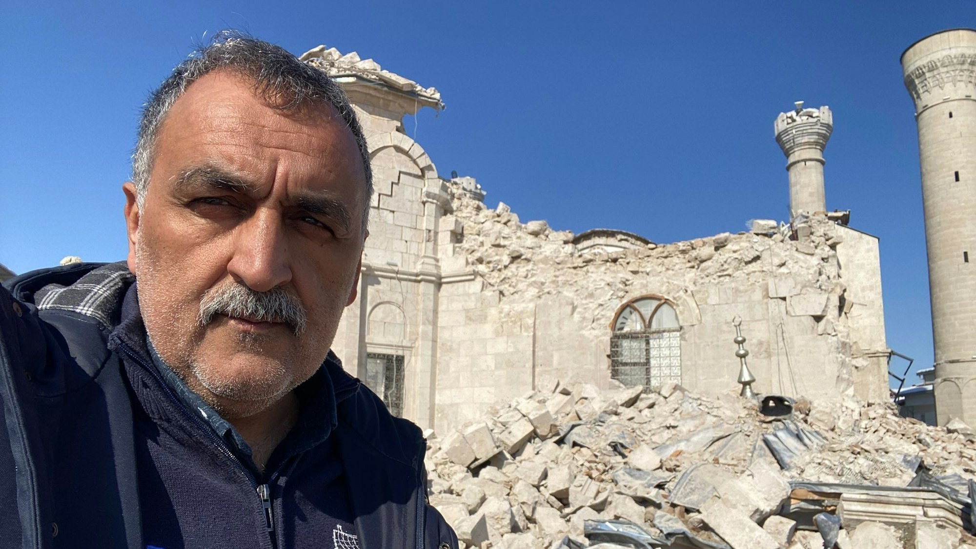 Im Bild ist ein Mann, der vor zerstörten Häusern steht.
