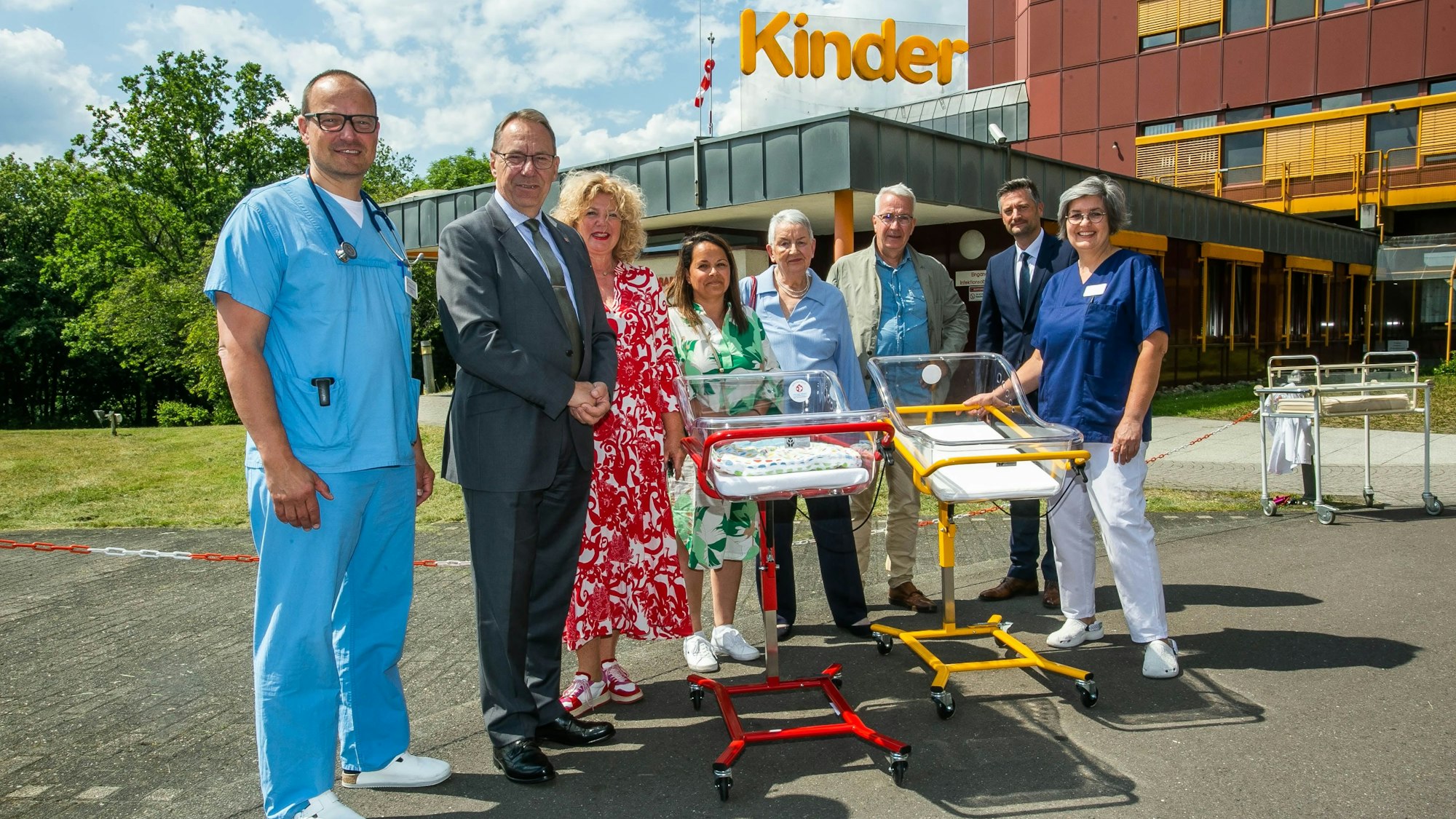 Mehrere Menschen, darunter auch Ärzte, stehen mit zwei neuen Kinderbetten vor dem Eingang einer Kinderklinik.