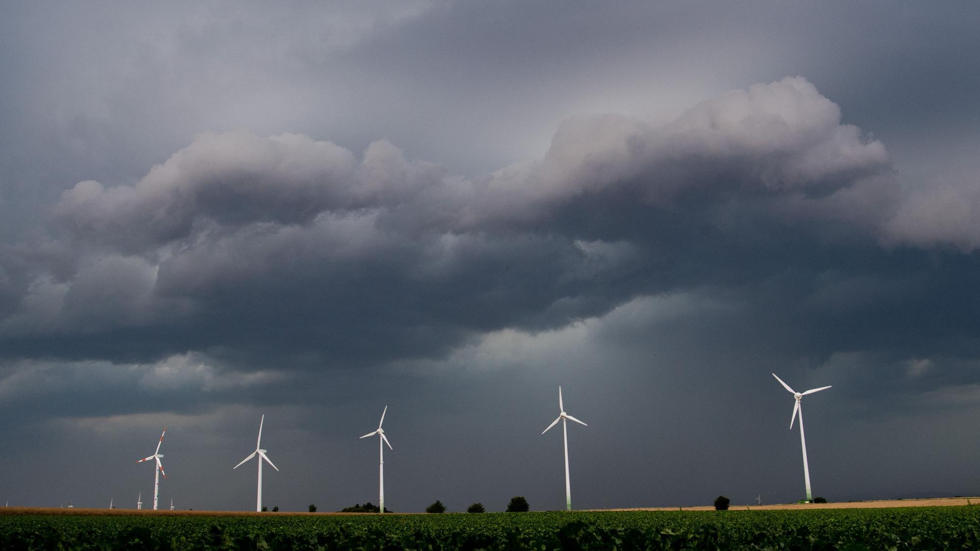 Eine Unwetterfront und dunkle Wolken ziehen über den Himmel. Davor ragen fünf Windräder in den Himmel.
