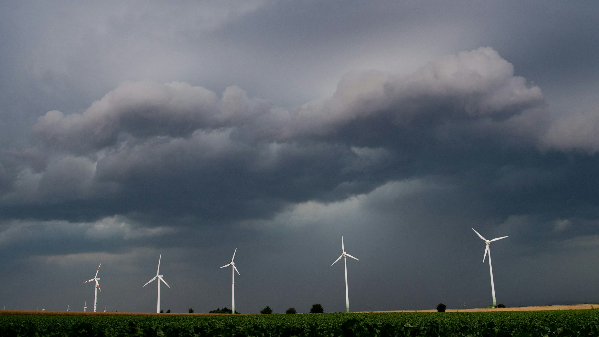 Eine Unwetterfront und dunkle Wolken ziehen über den Himmel. Davor ragen fünf Windräder in den Himmel.