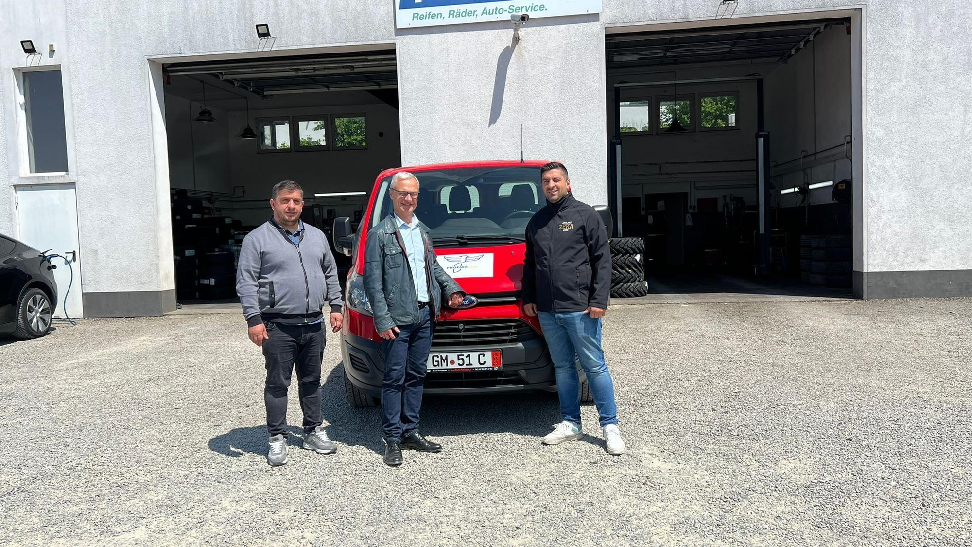 Mittig vor zwei geöffneten Garagentoren steht ein roter Lieferwagen. Vor diesem posieren drei Männer für die Kameraaufnahme.