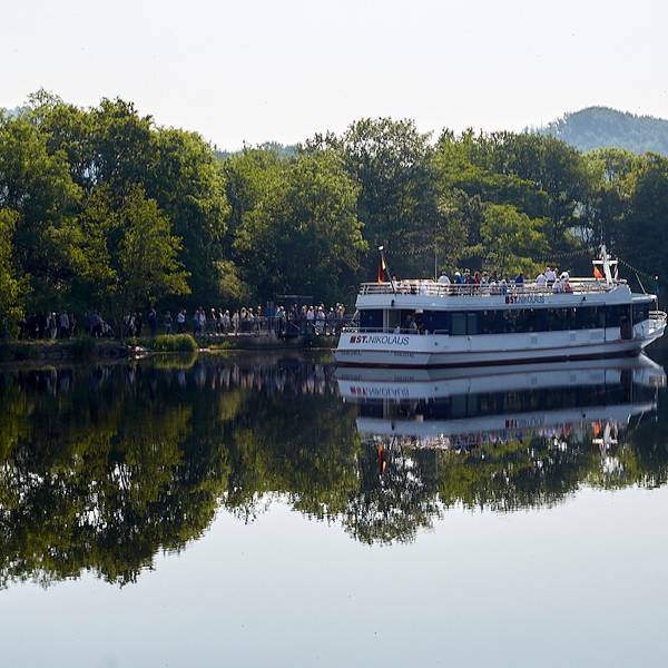 Bäume spiegeln sich im klaren Wasser des Rursees. Ein Schiff liegt am Steg, eine Schlange Menschen steht davor, um an Bord zu gehen.