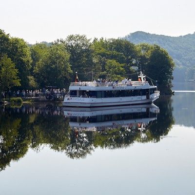 Bäume spiegeln sich im klaren Wasser des Rursees. Ein Schiff liegt am Steg, eine Schlange Menschen steht davor, um an Bord zu gehen.