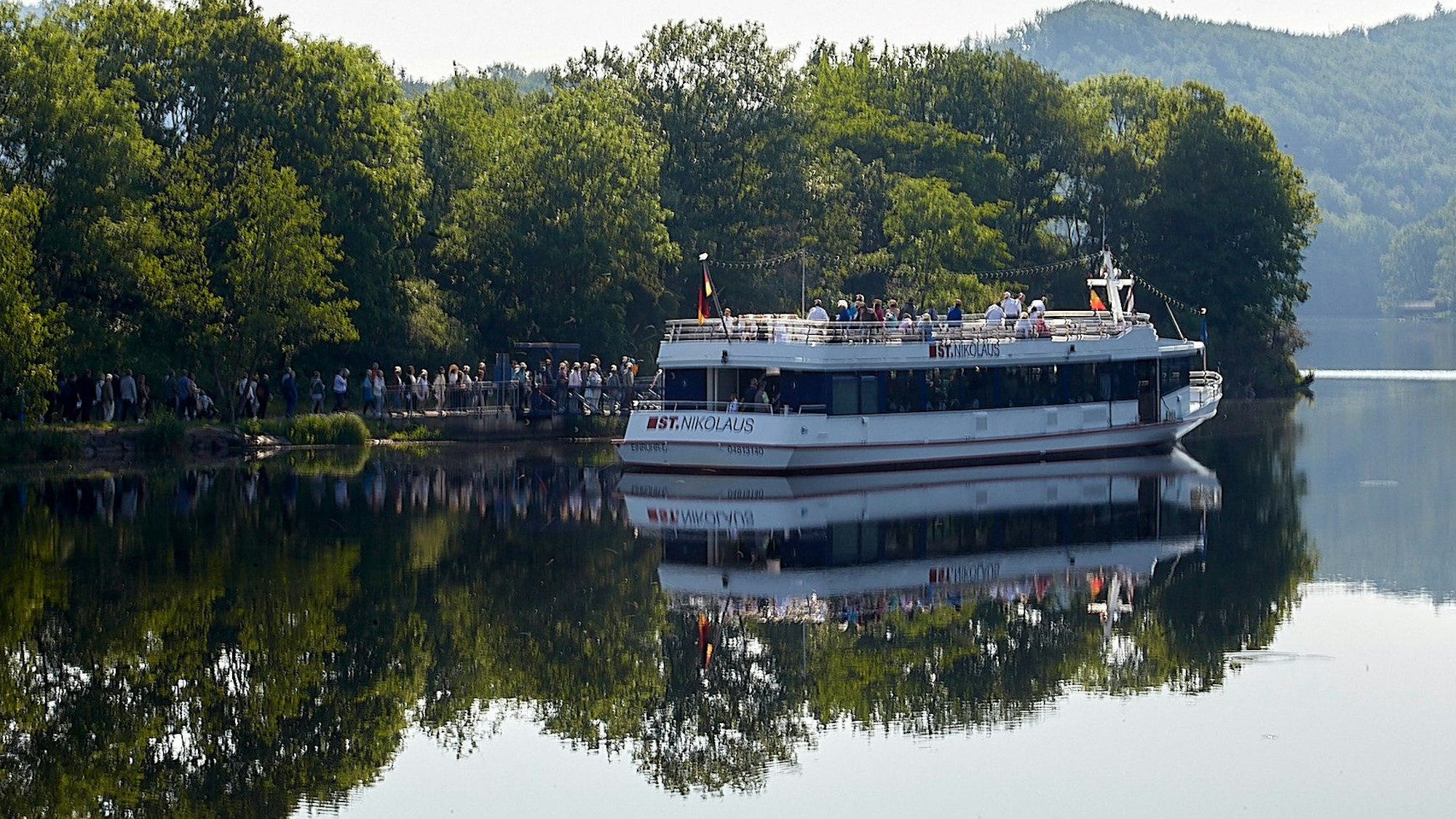 Bäume spiegeln sich im klaren Wasser des Rursees. Ein Schiff liegt am Steg, eine Schlange Menschen steht davor, um an Bord zu gehen.