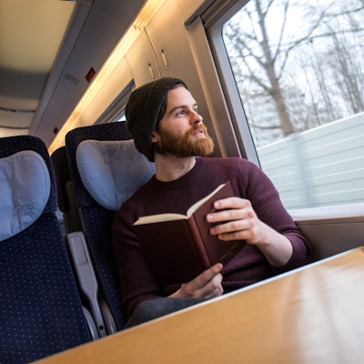 Ein junger Mann sitzt an einem Tisch im ICE. Er hält ein Buch in der Hand und schaut aus dem Fenster.