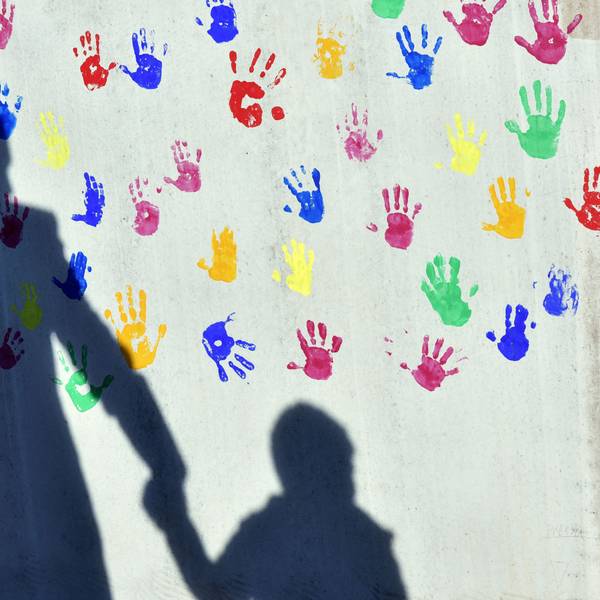 Ein Mann mit einem Kind auf dem Arm und einem an der Hand wirft einen Schatten auf eine mit bunten Handabdrücken bemalte Wand einer Kindertagesstätte.