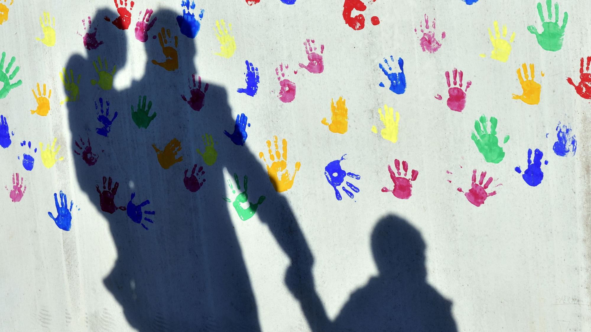 Ein Mann mit einem Kind auf dem Arm und einem an der Hand wirft einen Schatten auf eine mit bunten Handabdrücken bemalte Wand einer Kindertagesstätte.