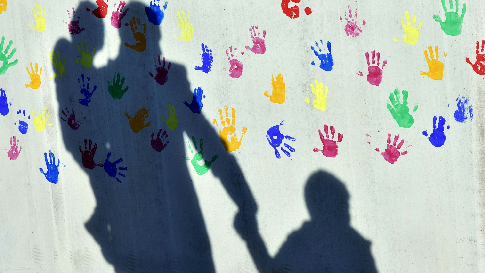 Ein Mann mit einem Kind auf dem Arm und einem an der Hand wirft einen Schatten auf eine mit bunten Handabdrücken bemalte Wand einer Kindertagesstätte.