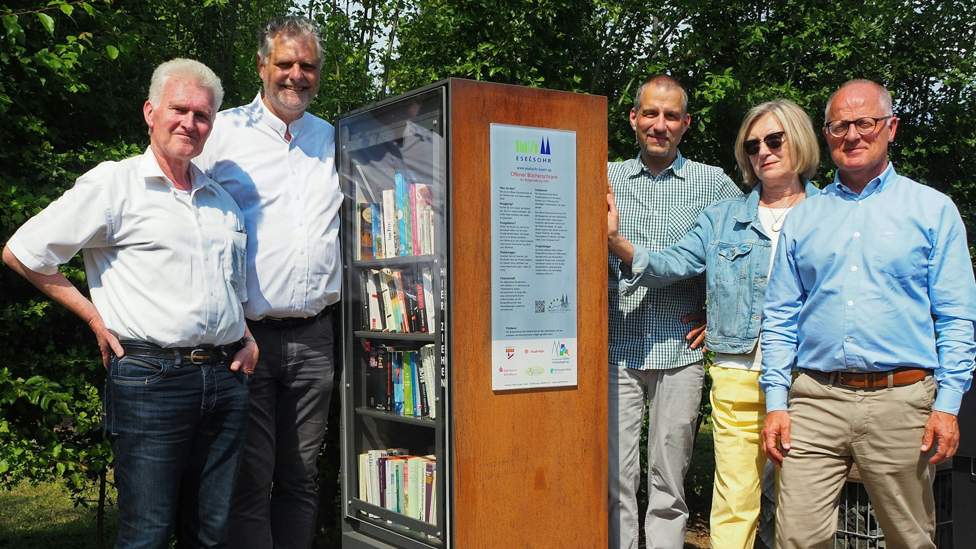 Vier Männer und eine Frau stehen links und rechts von einem Bücherschrank, an dem eine Tafel angebracht ist.