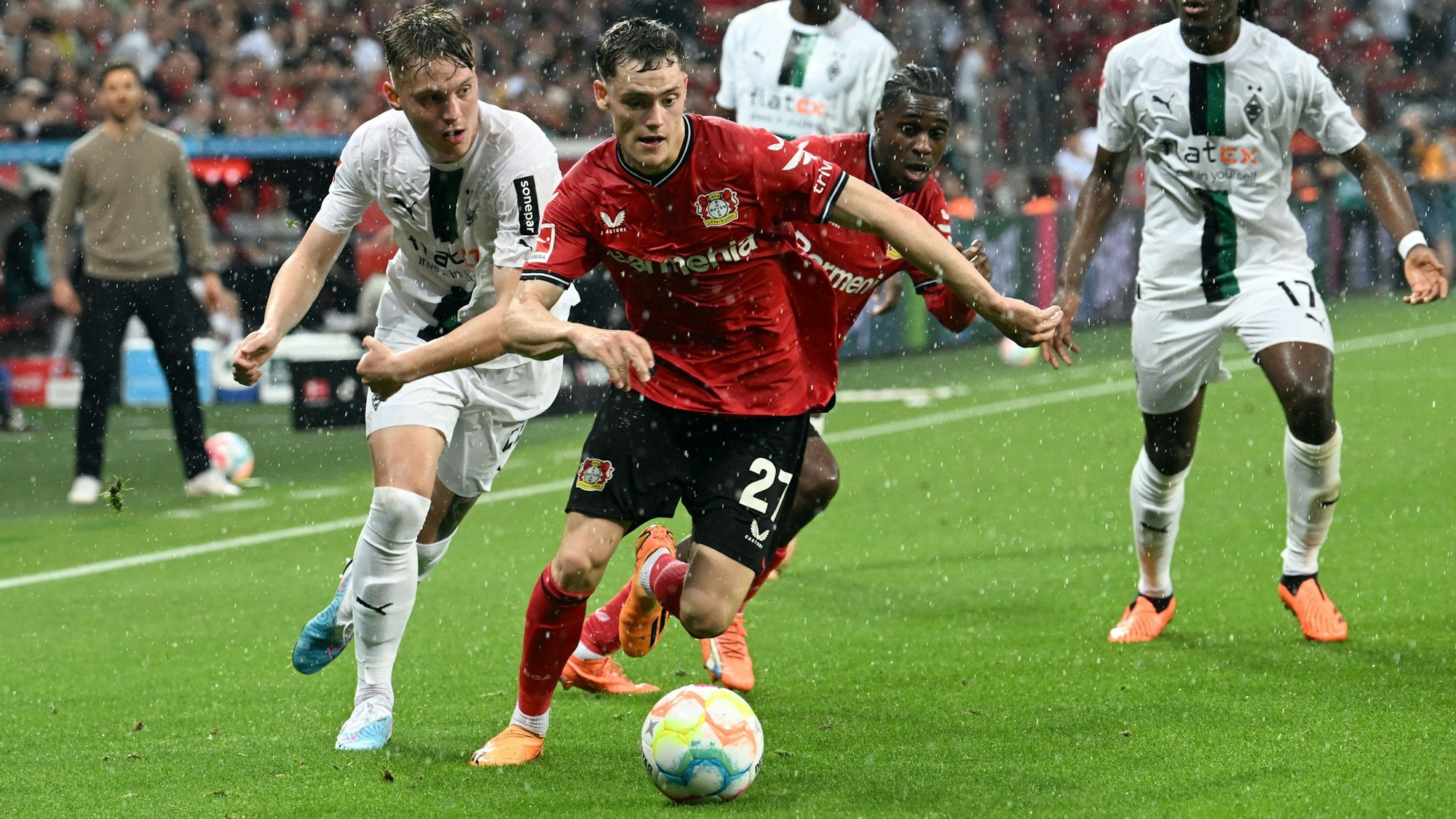 Leverkusens Florian Wirtz und Mönchengladbachs Luca Netz kämpfen um den Fußball. Es regnet im Stadion. Im Hintergrund sind weitere Fußballspieler sowie der Trainer Xabi Alonso zu sehen.