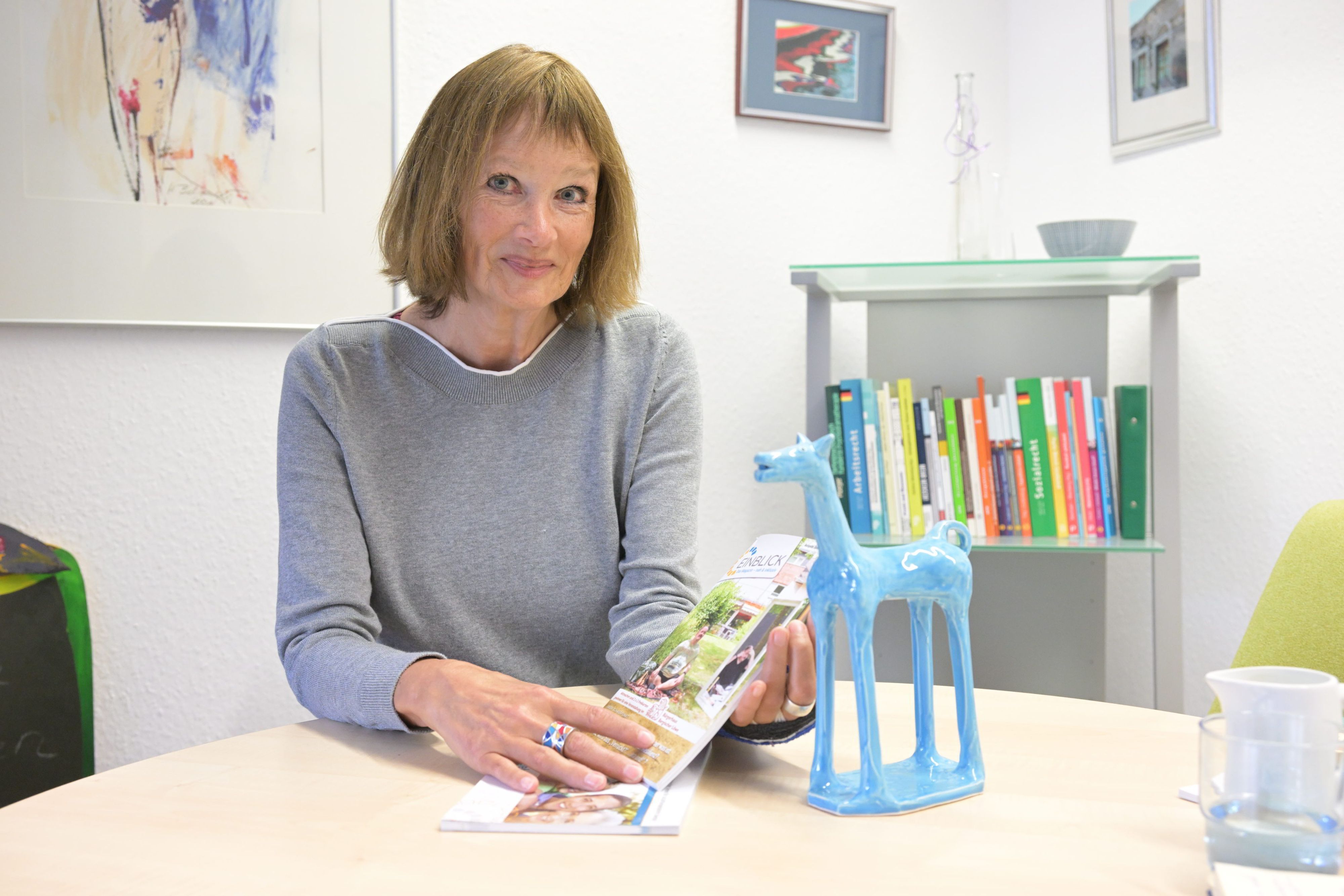 Porträt Claudia Seydholdt, sie sitzt am Tisch und blickt in die Kamera. Neben ihr steht eine blaue Pferde-Figur.