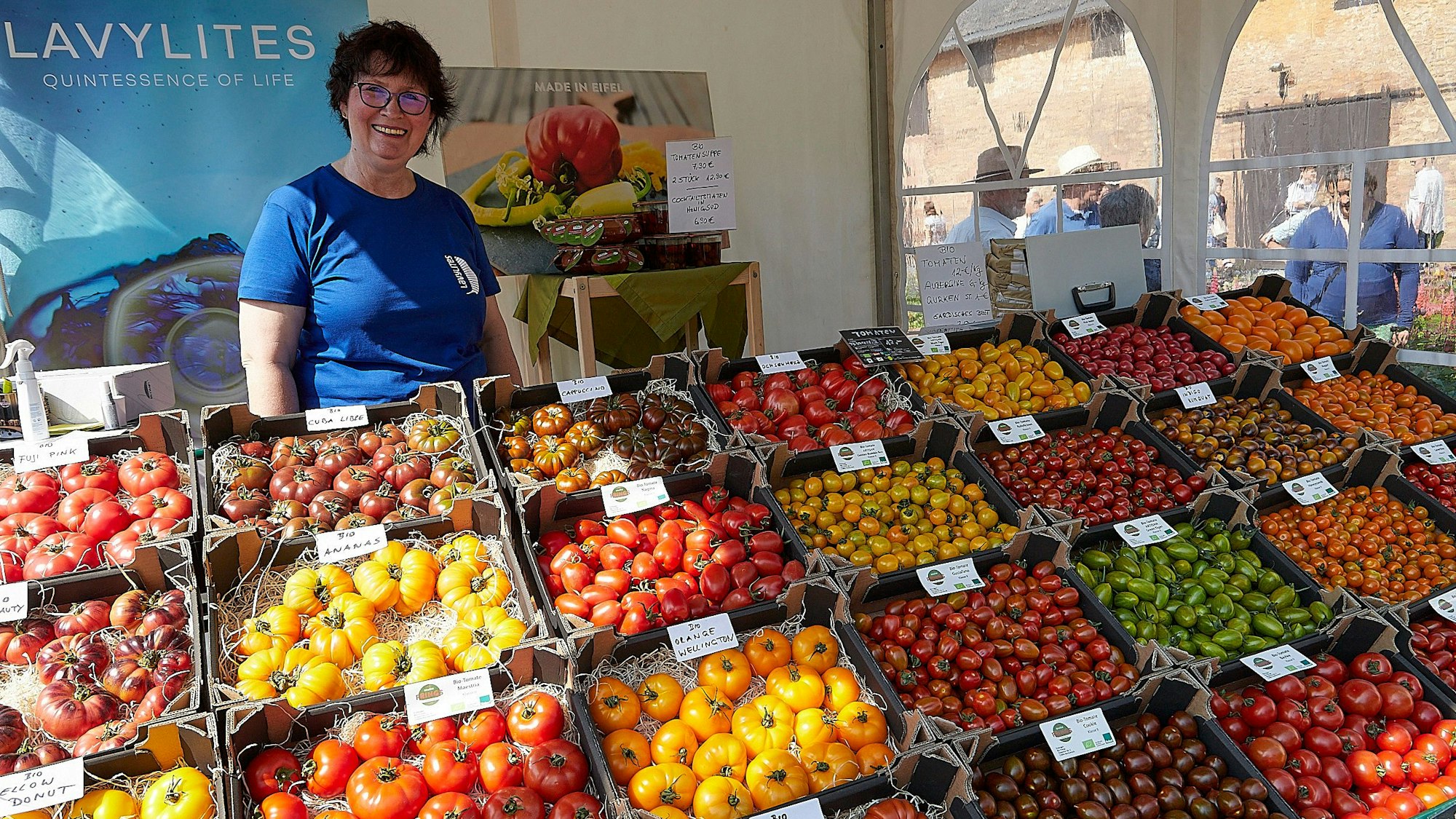 Auf dem Bild ist ein Marktstand mit zahlreichen Kisten zu sehen, in denen verschiedene Tomatensorten angeboten werden, dahinter die Verkäuferin.