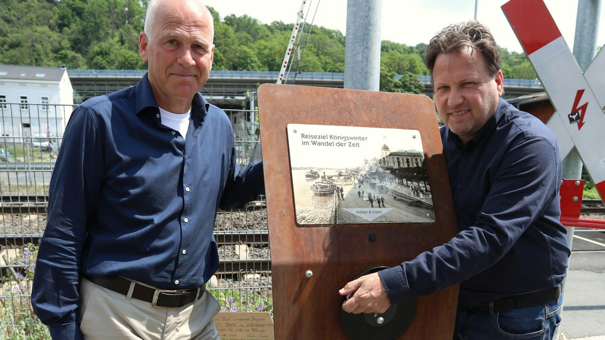 Zwei Männer stehen neben der neuen Hörstation, die aus Cortenstahl gefertigt wurde. Hinter ihnen verläuft die Bahntrasse.