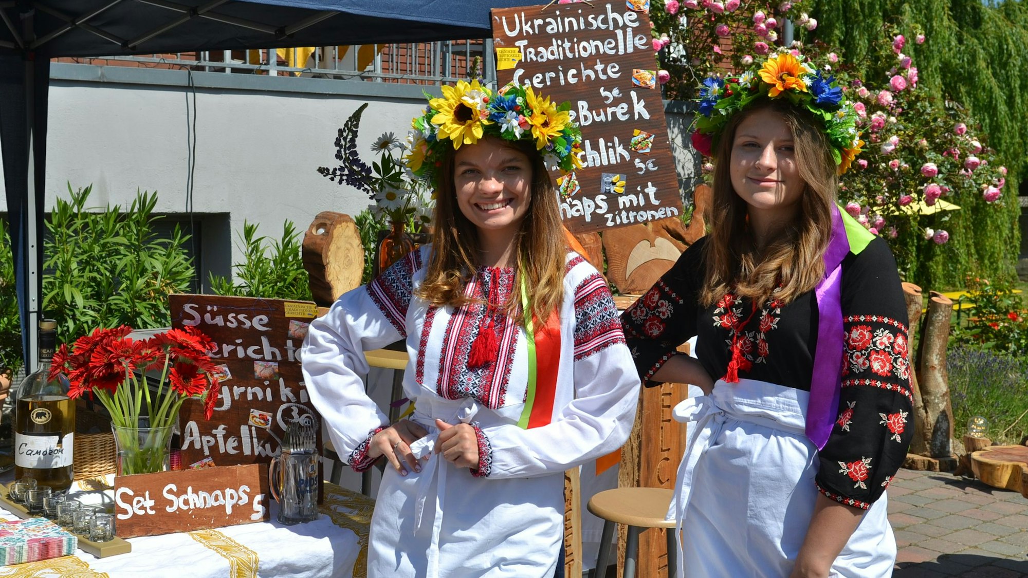 Zwei junge Frauen mit einem bunten Blumenkranz auf dem Kopf stehen vor einem Verkaufsstand auf dem Hoffest des Kulturhofs Velbrück in Metternich.