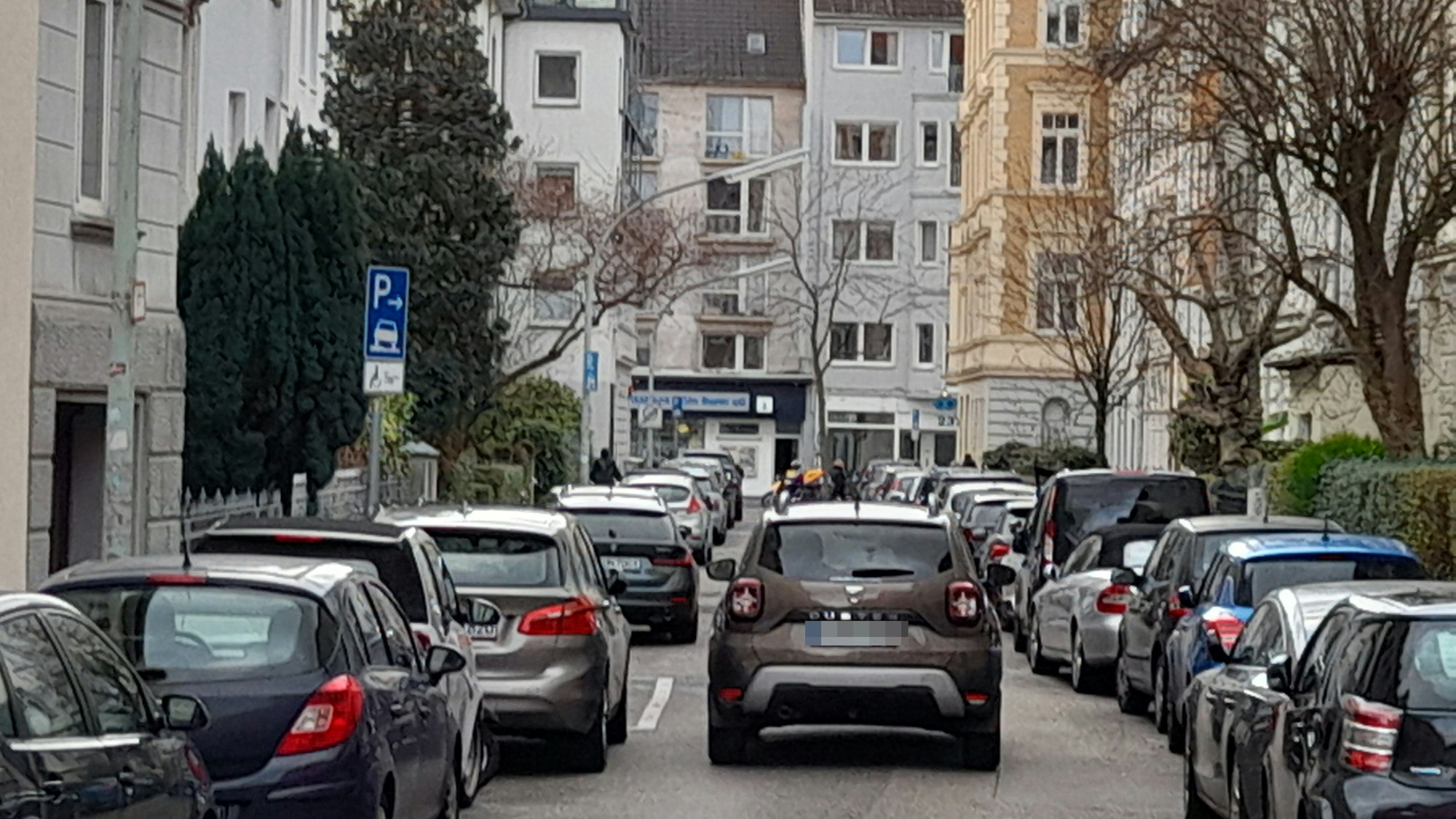 Ein Auto fährt durch eine schmale Straße, an der links und rechts Autos dicht an dicht parken.
