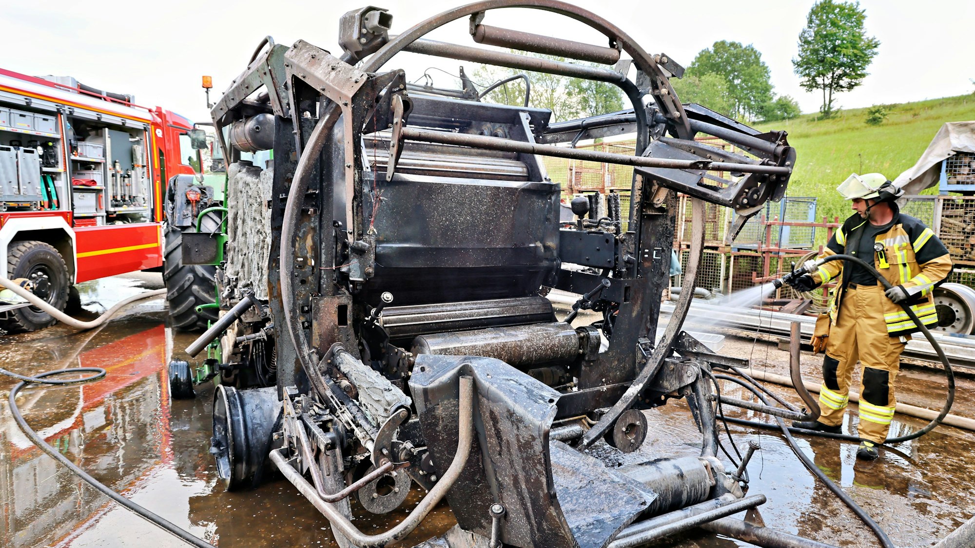 Ein Feuewrwehrmann kühlt mit einem Wasserschlauch das Gerippe der völlig zerstörten Rundballenpresse, die ins Freie geschleppt wurde.