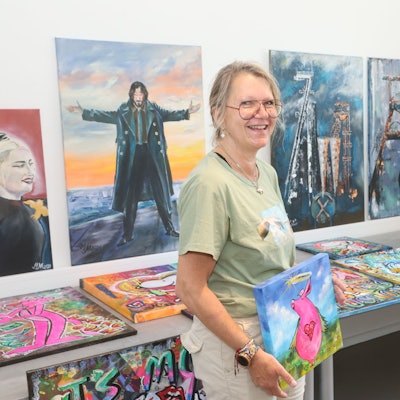 Eine Frau steht vor einer Bilderausstellung. Sie selbst hält ein Bild in der Hand, auf dem ein pinkes Schwein mit Herzkette um den Hals zu sehen ist.