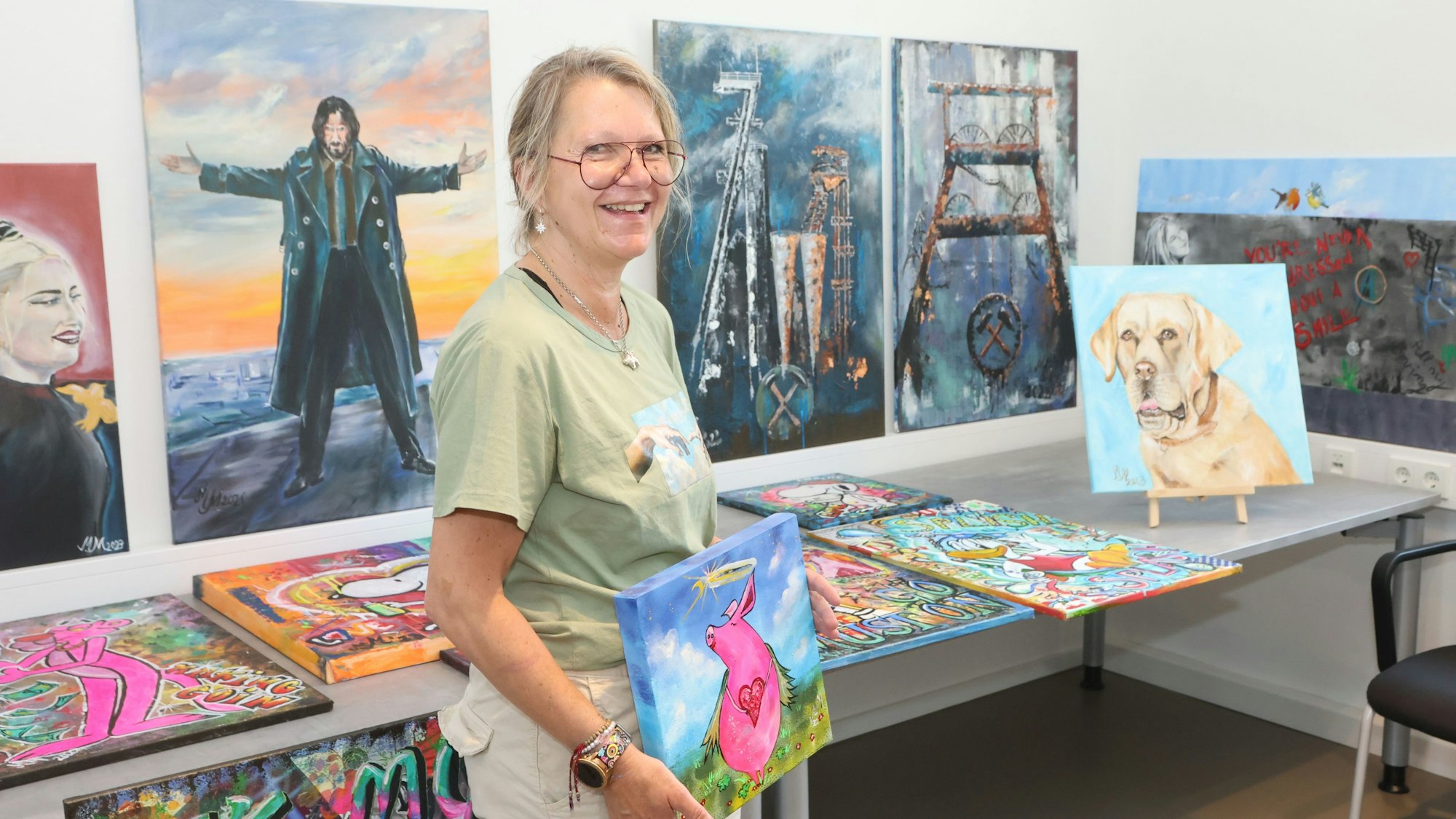 Eine Frau steht vor einer Bilderausstellung. Sie selbst hält ein Bild in der Hand, auf dem ein pinkes Schwein mit Herzkette um den Hals zu sehen ist.