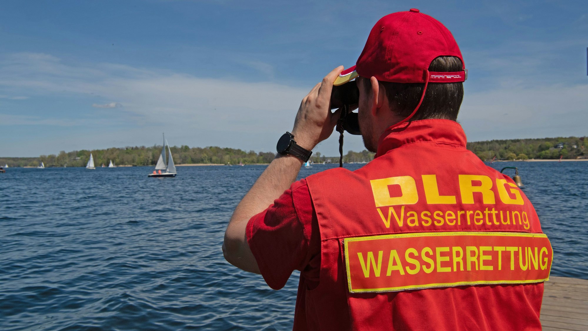 Ein Mann mit einer DLRG-Weste mit der Aufschrift Wasserrettung schaut durch ein Fernglas auf ein Gewässer.