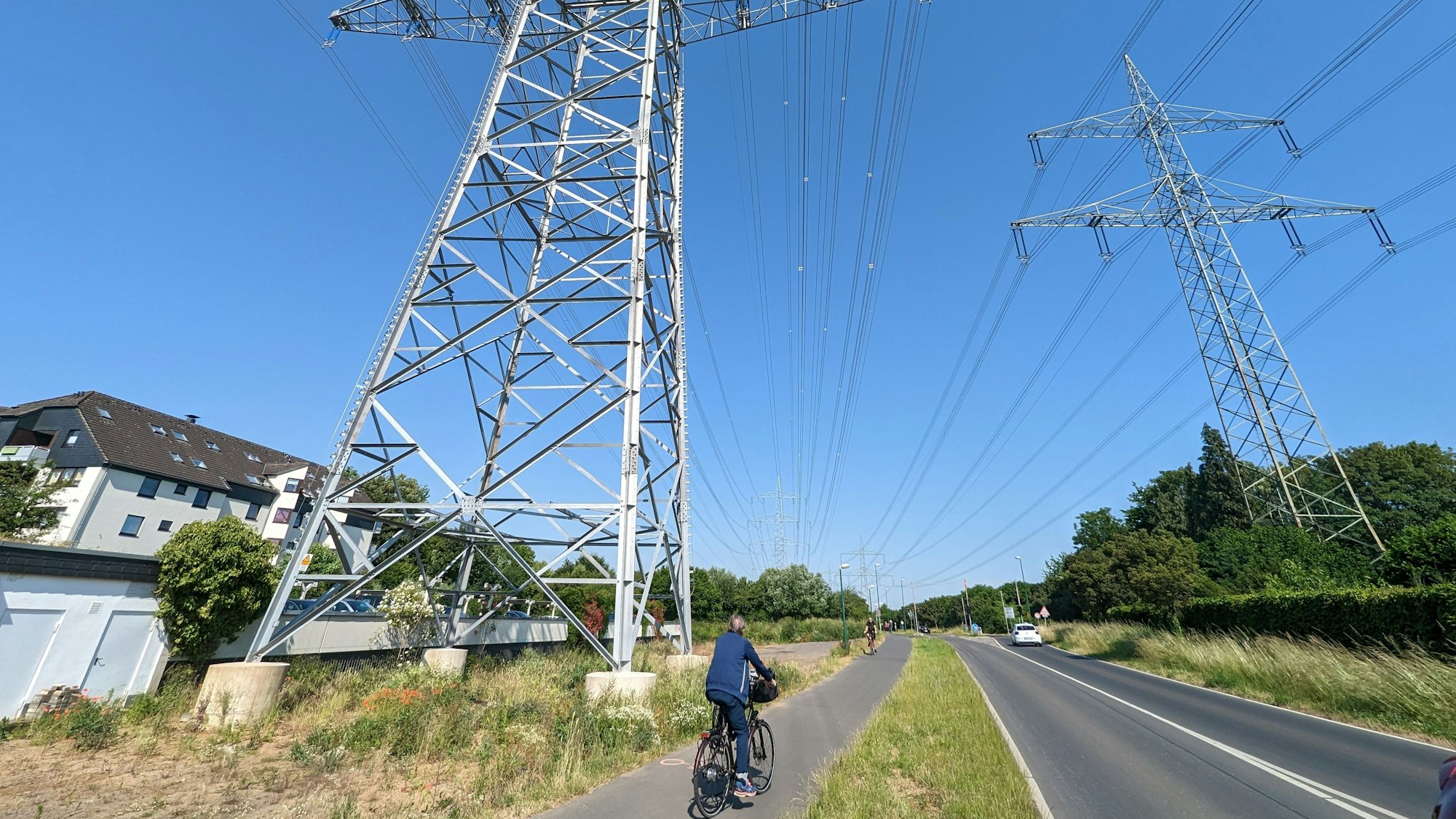 Das Foto zeigt den neuen Hochspannungsmasten an der Straße in den Höhnen, unmittelbar daneben ist Wohnbebauung. Radfahrer passieren den Radweg.
