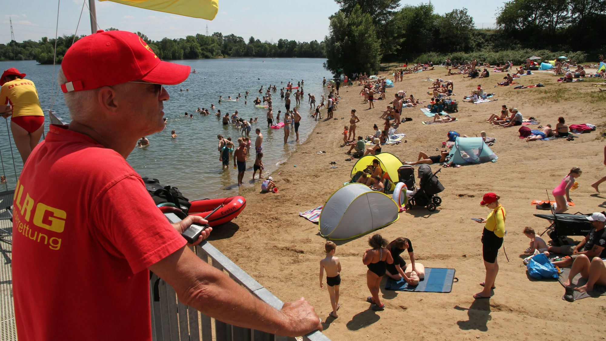 Ein Mitglied der DLRG steht auf einem Ausguck. Er trägt ein rotes T-Shirt und eine rote Schirmmütze. An einem Mast weht die rot-gelbe Fahne der DLRG, am Strand und im Wasser sind viele Menschen.
