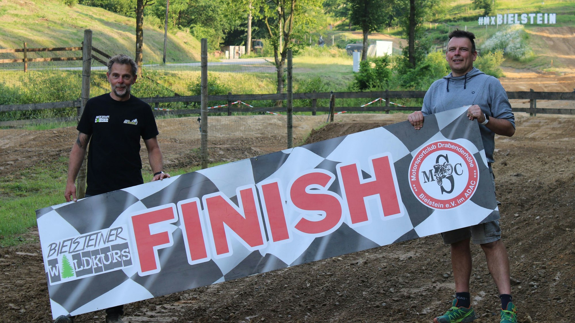 Zwei Männer stehen auf einem Waldkurs und halten ein Plakat in den Händen mit der Aufschrift „Bielsteiner Waldkurs – Finish“.