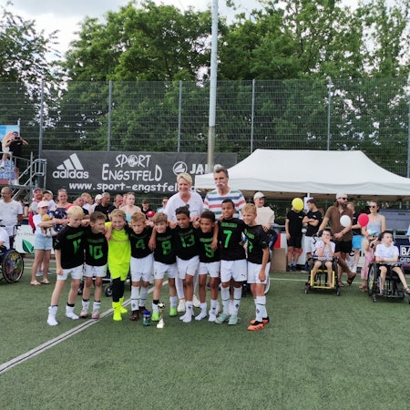 Eine Fußballmannschaft, bestehend aus Jungs, stehen in einer Reihe. Im Hintergrund ist Handballlegende Heiner Brand zu sehen.