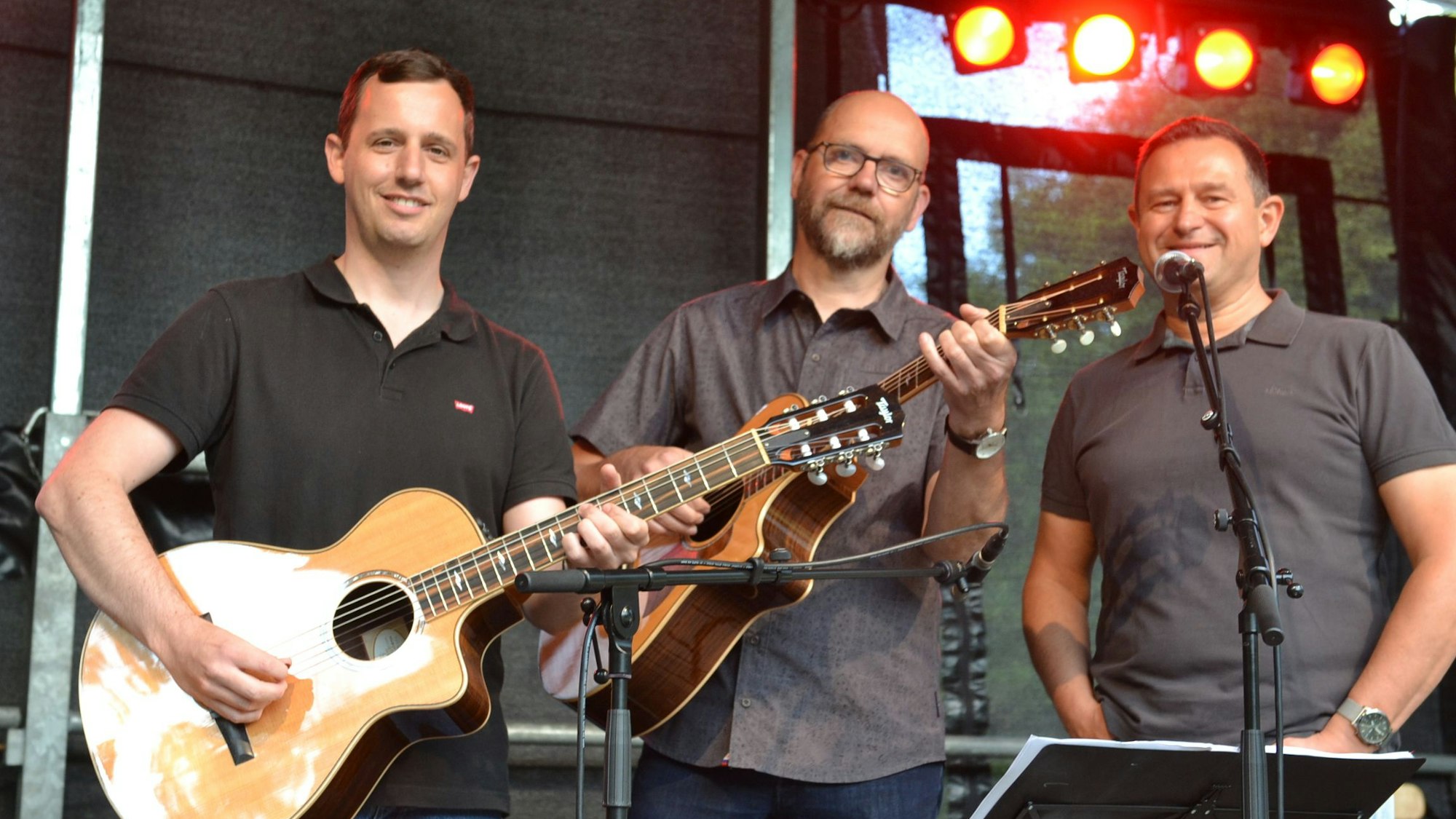 Zwei Männer mit Gitarre und ein Mann am Mikrofon stehen auf einer Bühne.