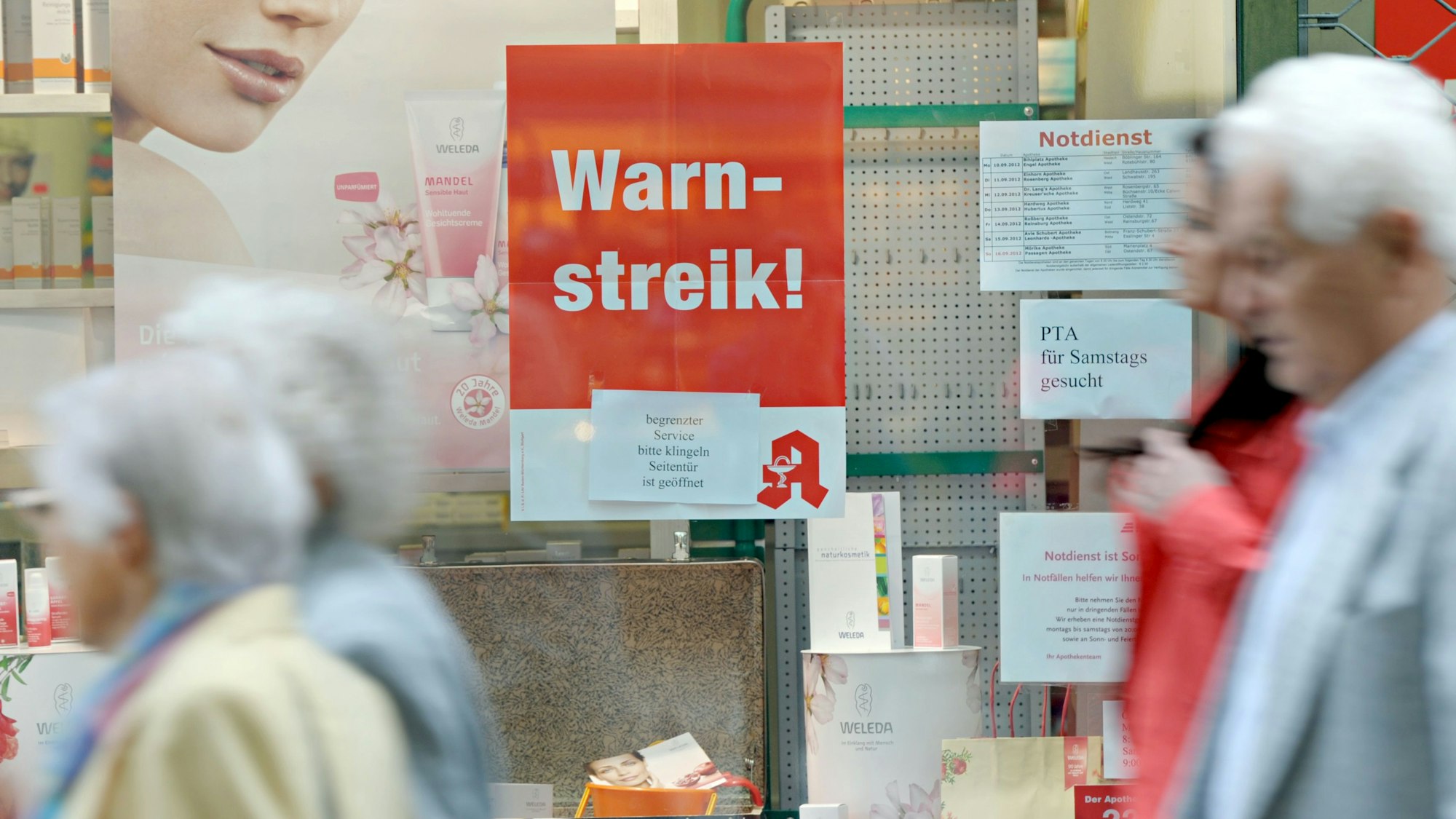 Passanten gehen an einem Schaufenster einer geschlossenen Apotheke vorbei, in dem ein Schild mit der Aufschrift „Warnstreik!“