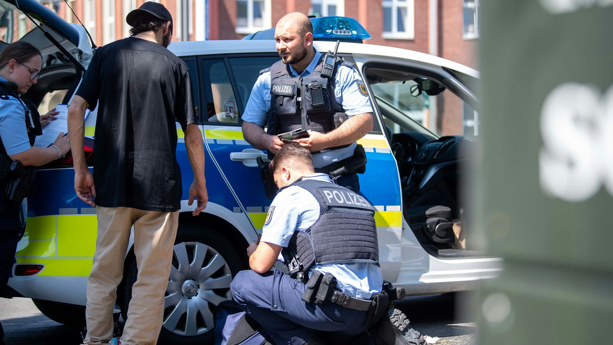 Ein Polizist steht vor einem Streifenwagen und spricht mit einem Mann. Ein zweiter Polizist kniet auf dem Boden und durchsucht eine Tasche.