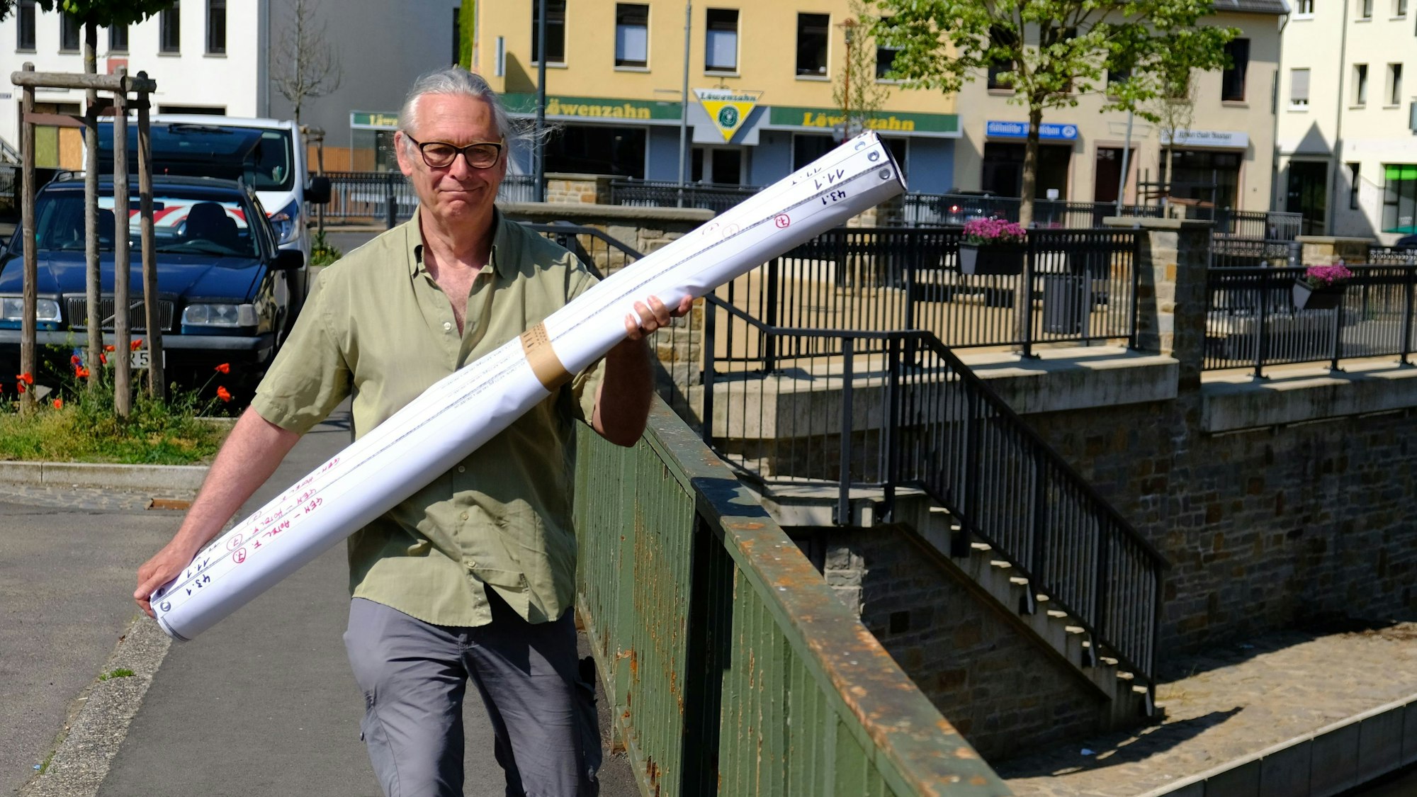 Kurator Jürgen A. Roder trägt einige gerollte Kunstdrucke über die Urftbrücke in Gemünd.
