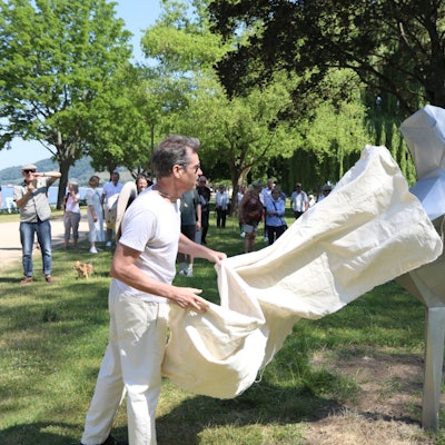 Ein Mann enthüllt ein etwa zwei Meter großes, silbernes Kunstwerk.Im Hintergrund stehende Menschen schauen zu.