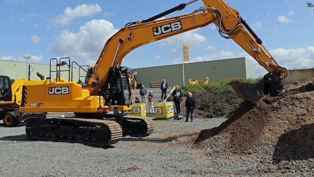 Ein gelber Bagger steht vor einigen Menschen auf einer Baustelle.